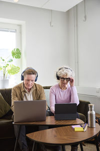 Senior man and woman sitting in living room and using laptop and digital tablet to edit podcast