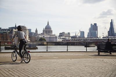 Woman riding bicycle on promenade by thames river against sky
