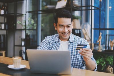 Smiling man holding credit card using laptop while sitting in cafe