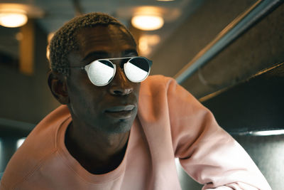 Adult african american man in pink sweatshirt and stylish sunglasses looking away while leaning on wall in illuminated tunnel