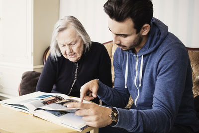 Young caretaker with senior woman reading book in living room at nursing home