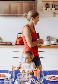 Boy having food by mother standing in kitchen at home