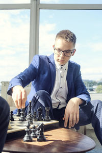 Dad and son are sitting at a table by a wide window and playing chess making moves with pieces