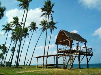 Thatched roof stilt hut by palm trees against sky