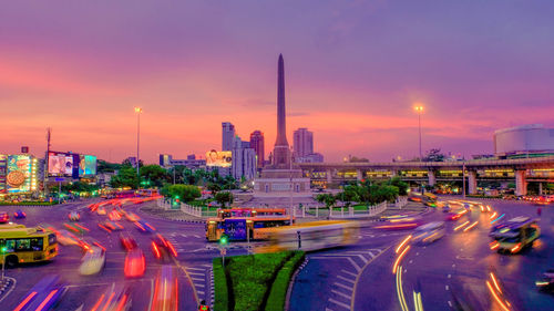 Light trails on city street by buildings against sky during sunset