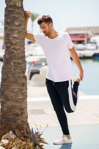 Full length of young man stretching by tree trunk at harbor