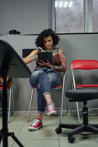 Concentrated tattoo master in casual clothes sitting on chair with tablet and pen while drawing tattoo sketches in light tattoo salon