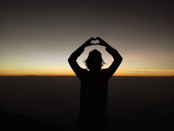 Silhouette man making heart shape with hands while standing against sky during sunset