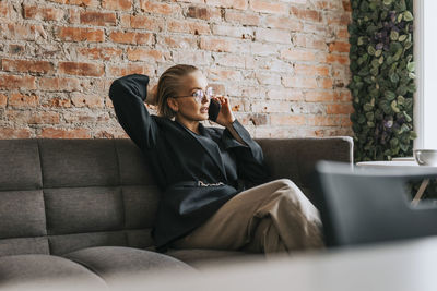 Young businesswoman with hand in hair talking on smart phone while sitting on sofa at office