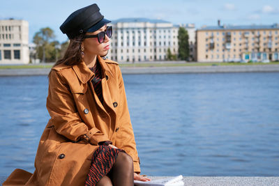 Young woman in sunglasses posing on embankment in brown coat on spring sunny day