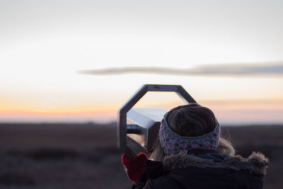 Rear view of young woman looking through coin-operated binoculars at beach during sunset