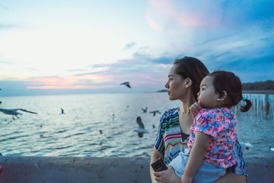 Mother carrying daughter while standing by sea against sky during sunset