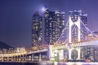 Illuminated bridge over river against buildings at night
