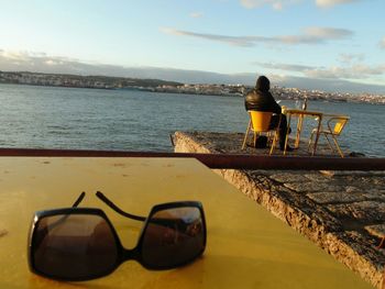 Sunglass on table with rear view of man sitting on chair by tagus river