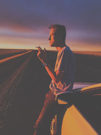 Side view of man smoking cigarette while standing by car on road against sky during sunset