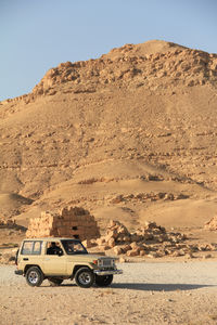 A rugged off-road vehicle travels through an expansive desert with majestic mountains and a blue sky