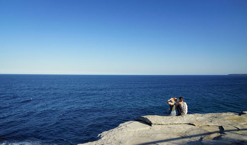 Couple sitting on rock by sea against clear blue sky