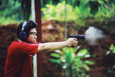 Side view of young man aiming handgun while standing against plants