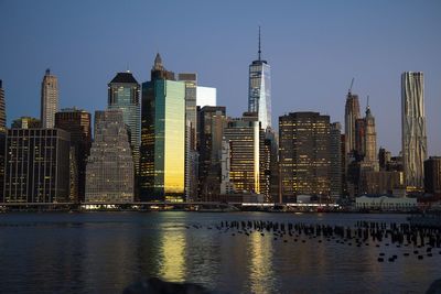 Modern skyscrapers by river against clear blue sky during sunset