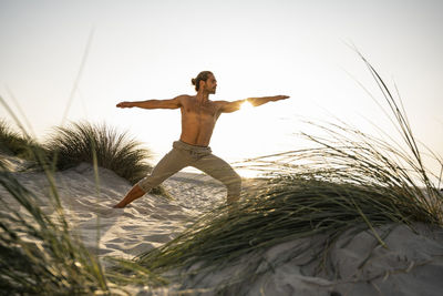 Shirtless young man practicing warrior 2 position yoga at beach against clear sky during sunset