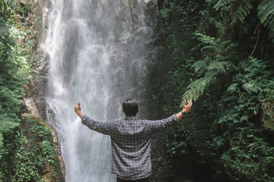 Rear view of man with arms outstretched looking at waterfall in forest