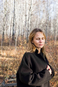 A young woman stands pensively in a dense forest. she is wearing a long black coat. 