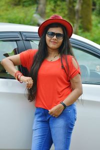 Portrait of young woman wearing sunglasses and hat while standing against car