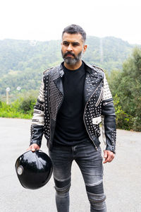 Serious brutal ethnic male biker in stylish leather jacket standing on asphalt road with helmet in hand near lush green trees growing in mountainous valley