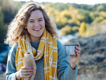 Young curly woman traveller smiling with coffee and hot dog in her hands at mountain in autumn