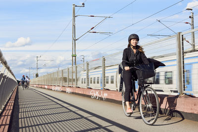 Mature woman cycling on road by fence against sky