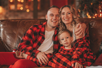 Smile active parents with small son in red checkered sleepwears waiting santa indoor.