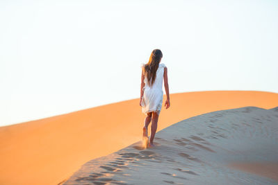 Rear view of girl walking on sand dunes at desert against clear sky
