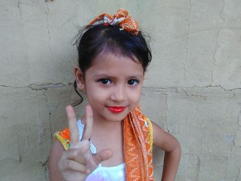 Close-up portrait of cute smiling girl showing peace sign against wall