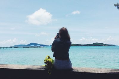 Rear view of woman sitting on retaining wall photographing sea against sky