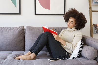 Woman writing in book while listening music through smart phone on sofa