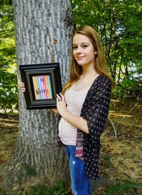Portrait of pregnant teenage girl holding frame while standing against tree trunk