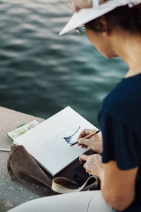 Cropped image of woman painting on paper while resting at retaining wall by sea