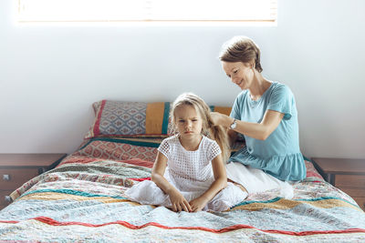 Mother tying daughter hair while sitting on bed at home