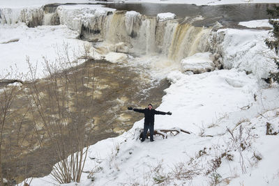 High angle view of man with arms outstretched standing on snow covered field by waterfall