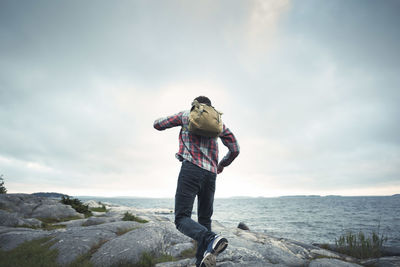 Rear view of wonderlust man jumping on rocks by sea against cloudy sky