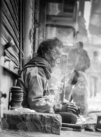 Shoeshiner working while sitting amidst smoke on sidewalk in city