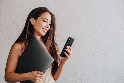 Young woman using phone while standing against wall