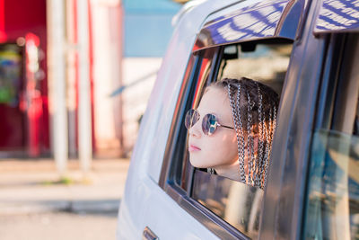 A cute teenage girl in sunglasses with afro-braids looks out the window while sitting in the car