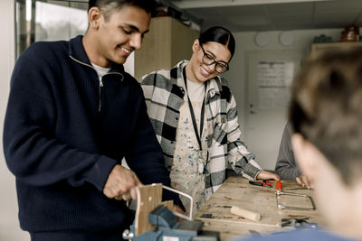 Happy female teacher standing with male student cutting wood during carpentry class