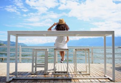 Rear view of woman sitting on stool at pier against sky