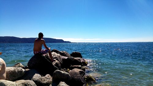 Man sitting on rock by sea against blue sky