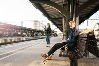 Young man sitting on bench with teenage girl in background at railroad station platform