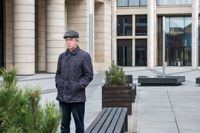 Adult caucasian man stands near modern building with columns. pensioner in seasonal clothes 