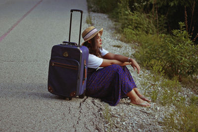 Teenage girl sitting by wheeled luggage on road