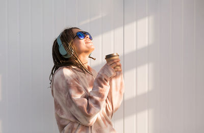 Dreadlock woman listening to music in headphones and drinking to go coffee, enjoying sunlight.
